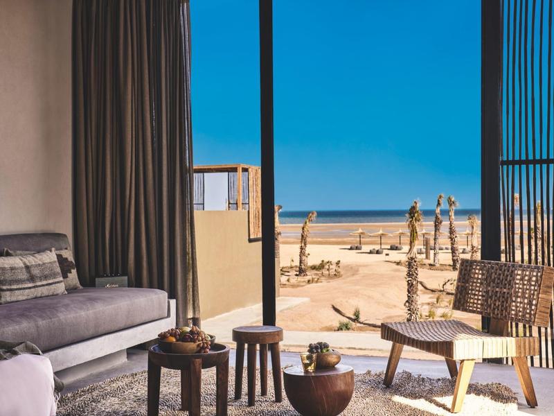 Helles Hotelzimmer mit Sitzmöbeln und Blick auf den Strand und das Meer bei klarem Himmel.