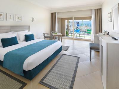 Helles Hotelzimmer mit großem Bett, türkisfarbener Bettdecke und Zugang zu Balkon mit Blick nach draußen.