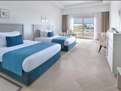 Helles Hotelzimmer mit zwei Einzelbetten, weißen Wänden, blau-weißer Bettwäsche und Balkonblick.