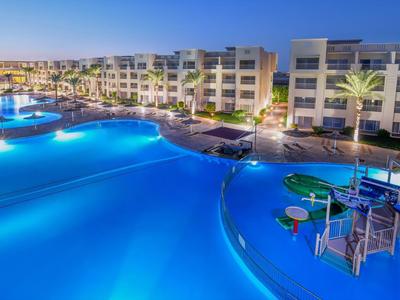 Großer Hotelpool mit blauer Beleuchtung, Palmen und modernen Gebäuden bei Abenddämmerung.