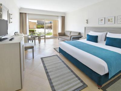 Helles Hotelzimmer mit großem Doppelbett, Sofa, Fernseher und Zugang zu Balkon mit Gartenblick.