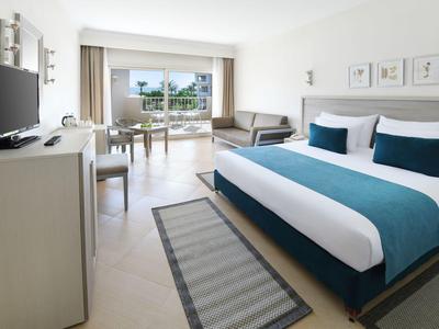 Helles Hotelzimmer mit großem Bett, blauem Bettlaken, Sofa, Schreibtisch, TV und Balkon mit Meerblick.