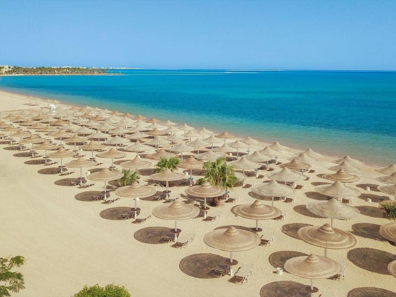 Langer Sandstrand mit vielen Strohhüten und ruhiger, blauer See im Hintergrund.