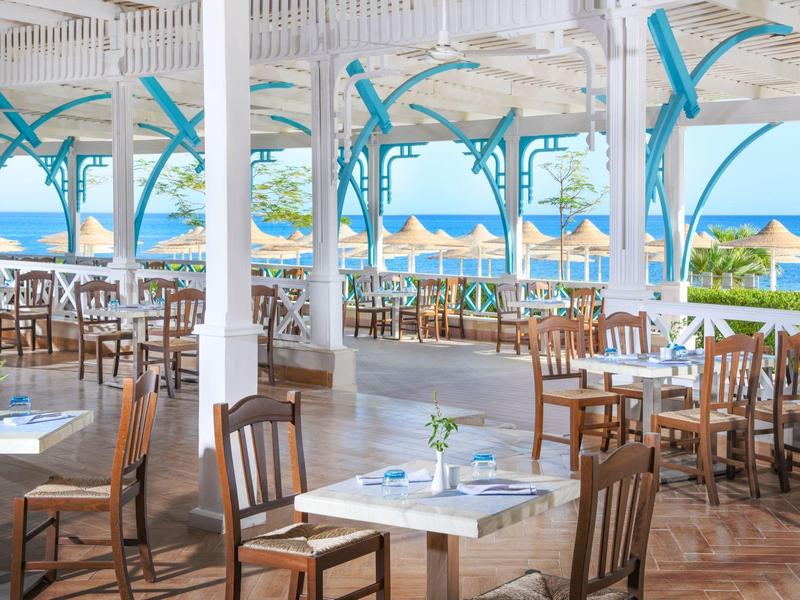 Offenes Restaurant mit Blick auf Strand und Meer, weiße Säulen, Holztische und -stühle.