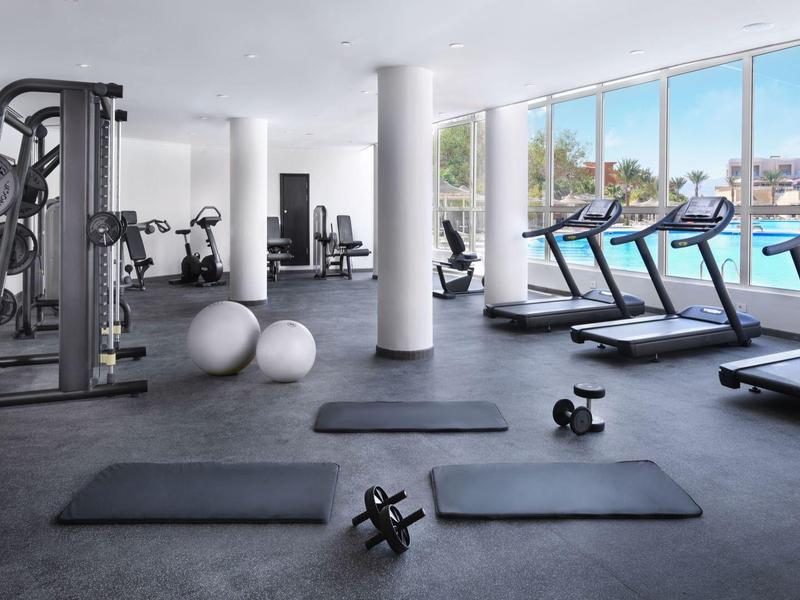Moderner Fitnessraum mit Geräten, Yogamatten und Blick auf den Pool durch große Fenster.