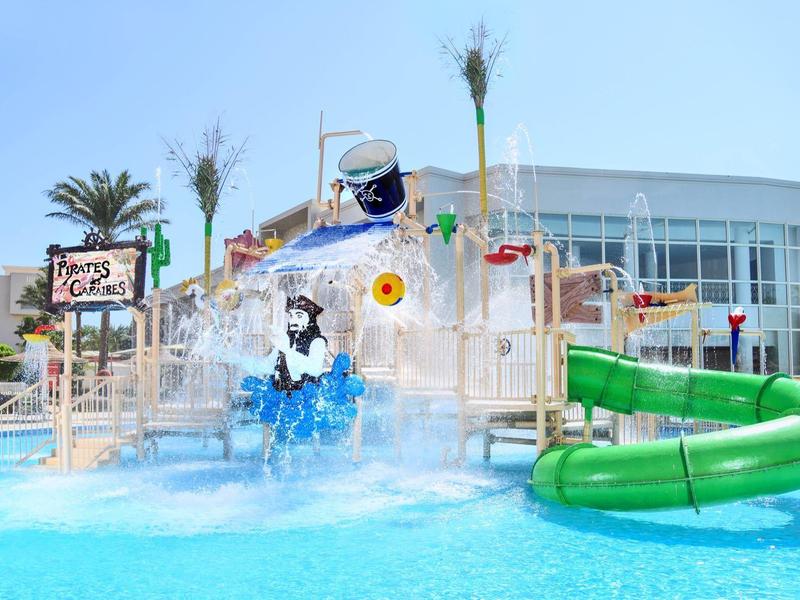 Bunter Wasserpark mit Rutsche und Sprinklern in einem Hotelpool unter blauem Himmel.