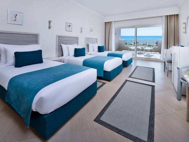 Helles Hotelzimmer mit drei Einzelbetten, blauen Akzenten und Meerblick vom Balkon.