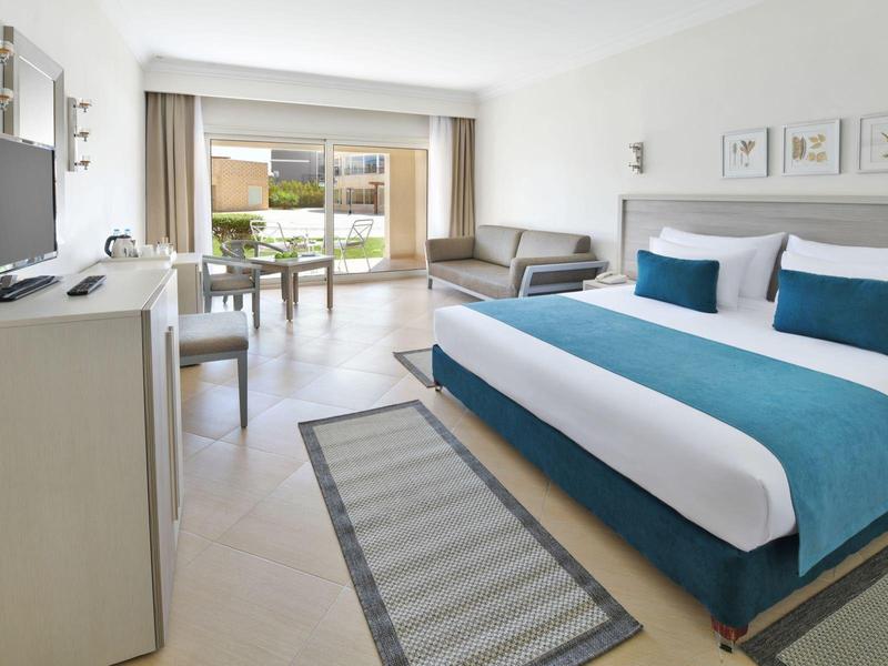 Helles Hotelzimmer mit großem Bett, Sitzbereich und Blick auf Terrasse und Garten.