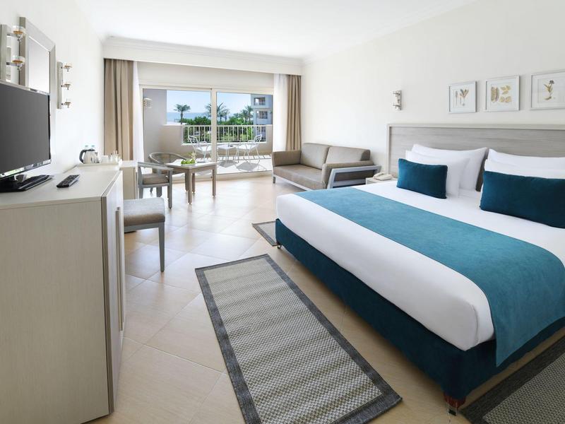 Helles Hotelzimmer mit Doppelbett, Fernseher, Sofa und Balkon mit Blick ins Grüne.