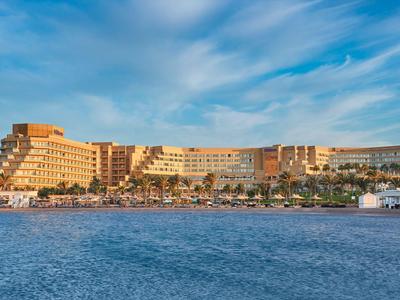 Large beachfront hotel resort under a cloudy sky with beach and palm trees.