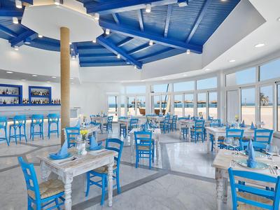 Bright restaurant with blue chairs, white tables, and large panoramic windows overlooking the sea.