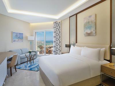 Helles Hotelzimmer mit großem Doppelbett, Schreibtisch, Fernseher und Balkon mit Meerblick.