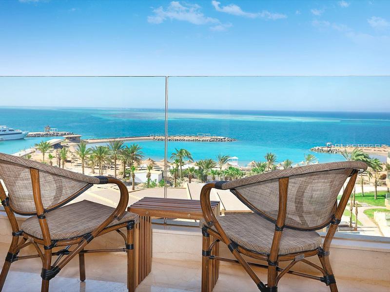 Balkon mit zwei Stühlen und kleinem Tisch, Blick aufs Meer, Strand, Palmen und Boote am Horizont.