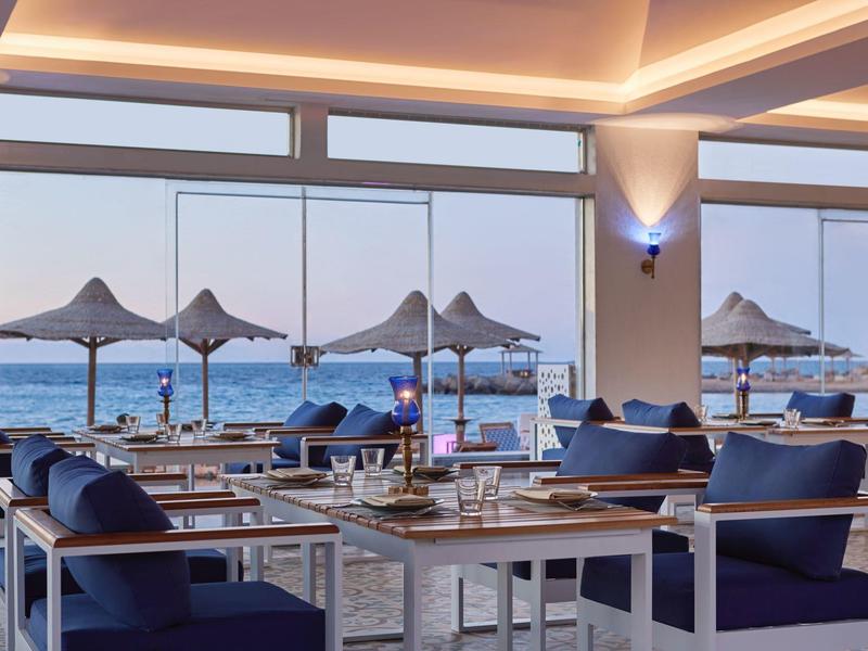 Modernes Restaurant mit blauen Sesseln und Tischen, große Fenster mit Meerblick und Sonnenschirmen draußen.