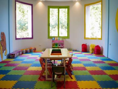 Bunter Kinderspielraum mit Tischen, Stühlen und großen Fenstern, bunte Schaumstoffmatten am Boden.