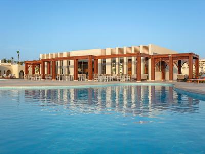 Großes Gebäude mit Säulen und Terrasse, davor großer Pool mit klar blauem Wasser bei sonnigem Himmel.