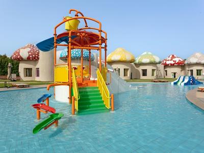 Buntes Wasserspiel mit Rutschen und Pilz-Häusern in flachem Pool unter blauem Himmel.