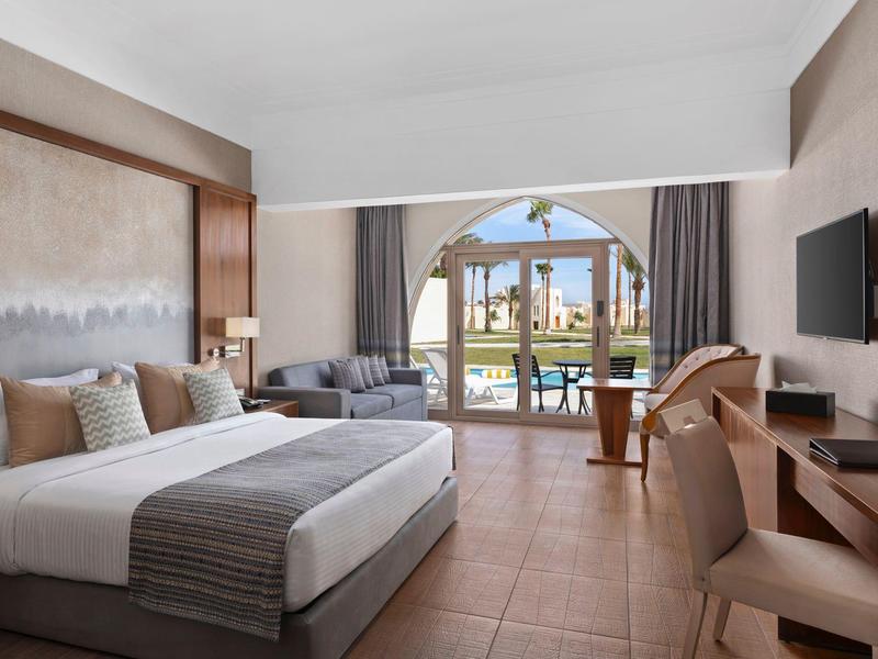 Modern hotel room with large bed, seating area, and view of a terrace with pool.