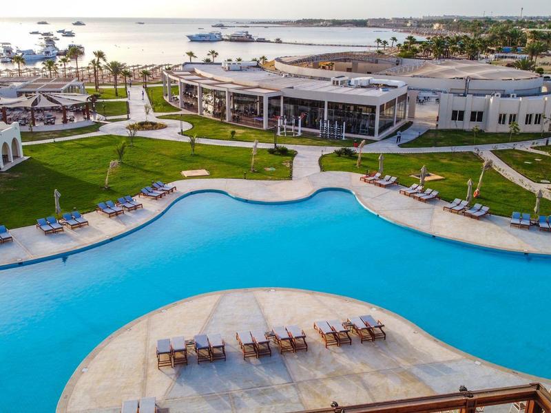 Large curved outdoor pool with sun loungers beside a modern hotel building by the sea.