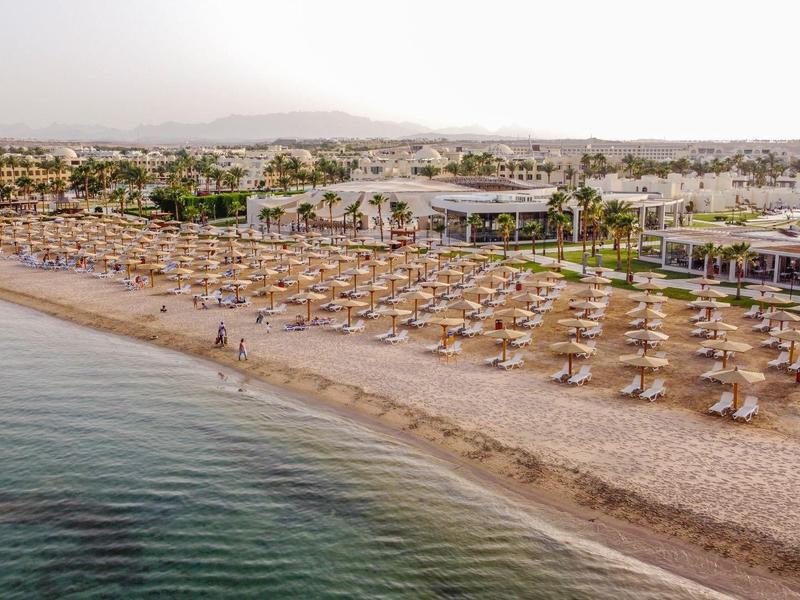 Wide sandy beach with numerous sun umbrellas and loungers by calm sea, hotel complex in the background.