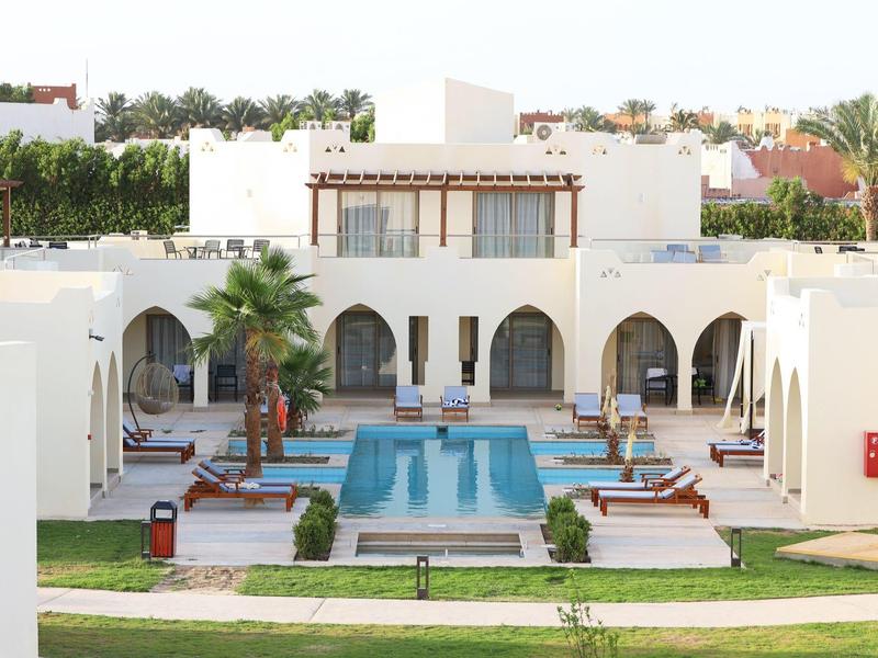 White hotel with pool, sun loungers, and green surroundings in sunny weather.