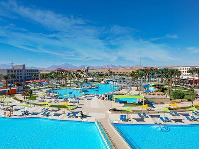 Großer Hotelpool mit Liegen, Wasserrutschen und Palmen unter blauem Himmel.