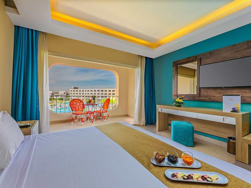 Helles Hotelzimmer mit blauem Akzent, Balkon mit Meerblick und bunten Stühlen, moderner TV-Wand.