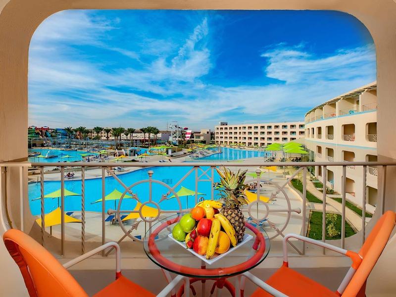 Balkon mit orangenen Stühlen, Obstkorb und Blick auf Pool, Hotelgebäude und blauen Himmel.