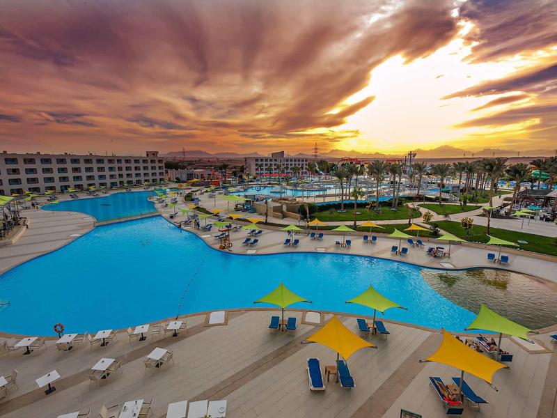 Großer, geschwungener Pool mit blauen Liegestühlen, gelben Sonnenschirmen und buntem Sonnenuntergang.