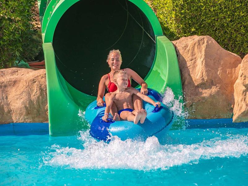 Zwei Kinder fahren fröhlich auf einer blauen Reifenrutsche in einen Pool mit klarem Wasser.