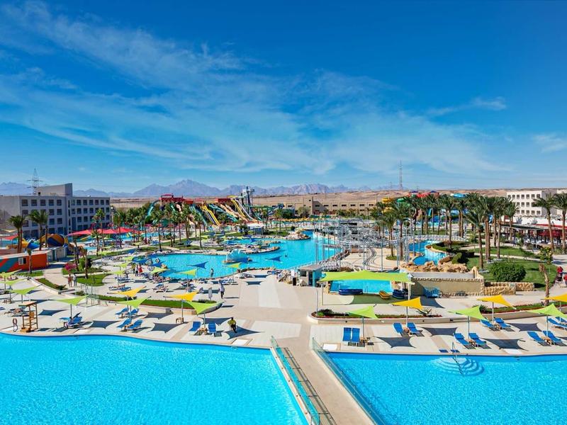 Großer Resort-Pool mit blauen Liegen, Sonnenschirmen und hügeliger Wüstenlandschaft im Hintergrund.
