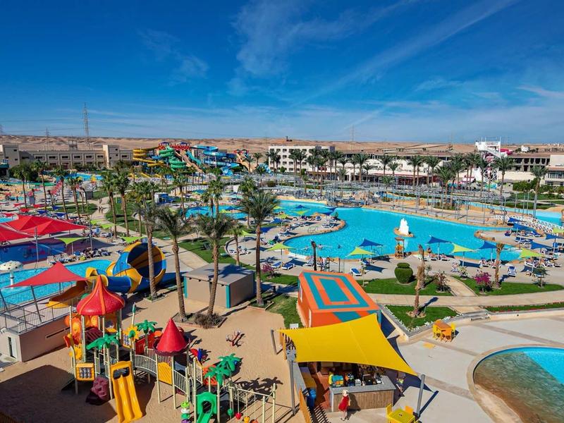 Großer Wasserpark mit mehreren Pools, bunten Sonnenschirmen, Spielplatz und Palmen unter blauem Himmel.