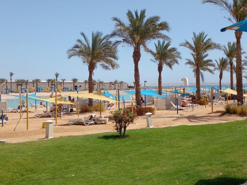 A resort with a large pool, palm trees, lounge chairs, and guests under clear skies.