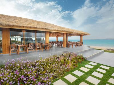 Restaurante abierto en la playa con techo de paja, mesas y sillas con vista al mar y flores al frente.