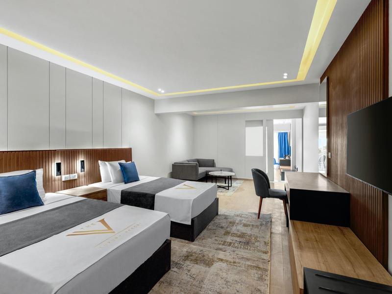 Moderne hotelkamer met twee bedden, zithoek, bureau en aan de muur gemonteerde tv, in neutrale kleuren.