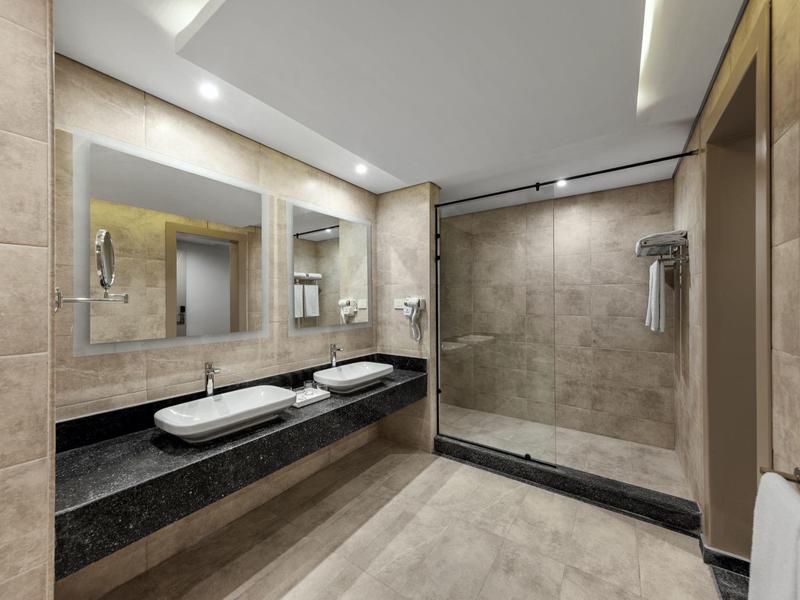 Moderne hotelbadkamer met dubbele wastafels, grote spiegel, inloopdouche en beige tegelwanden en vloer.