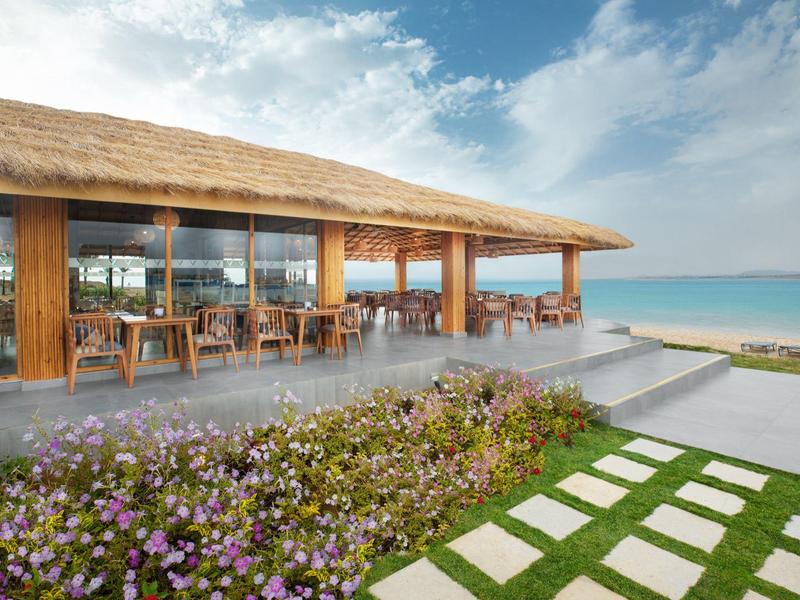 Strandrestaurant met houten stoelen, tafels onder een rieten dak, uitzicht op oceaan en bloementuin.