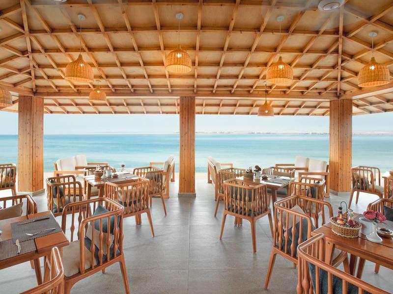 Openlucht restaurant met houten tafels en stoelen gericht op de oceaan onder een rieten dak.