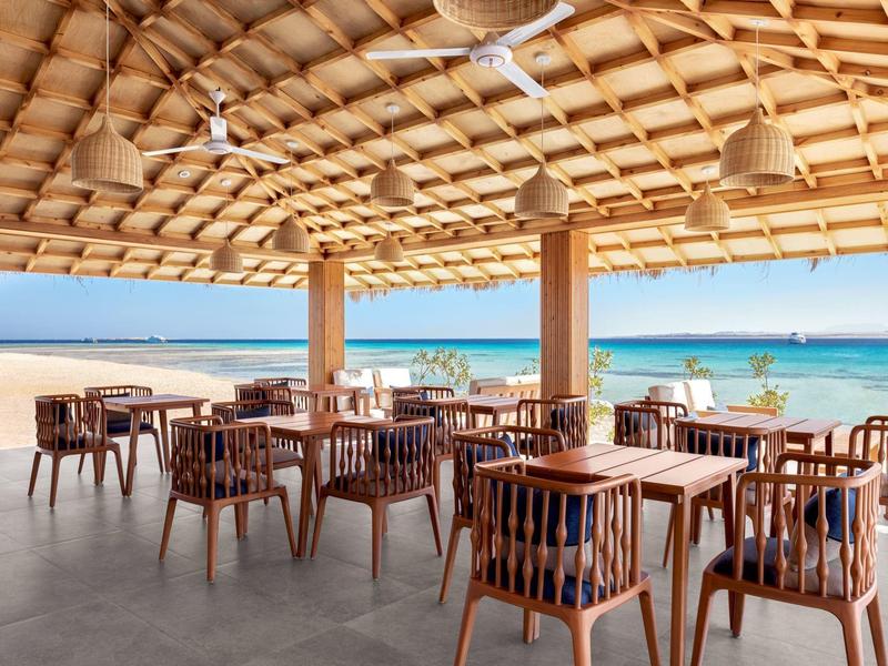 Buiteneetgedeelte met houten tafels en stoelen onder een rieten dak bij het strand.