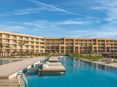 Complesso alberghiero moderno con grande piscina e lettini sotto un cielo sereno.