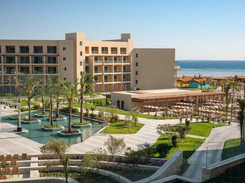 Complexe hôtelier avec piscine, palmiers et vue sur la mer sous un ciel clair.