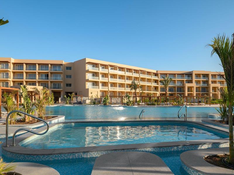Grand hôtel à plusieurs étages avec une grande piscine extérieure sous un ciel bleu clair.