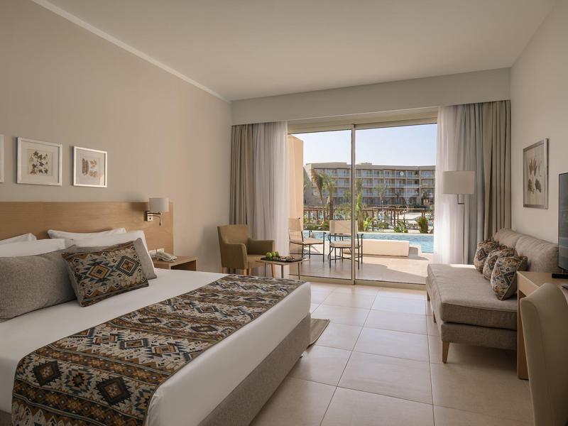 Chambre d'hôtel lumineuse avec lit double, coin salon et vue sur la piscine par de grandes portes coulissantes.