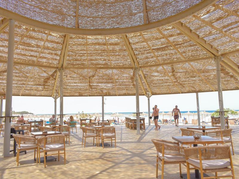 Offene Strandbar mit Holzstühlen und Tischen unter einem großen runden Strohdach bei Sonnenschein.