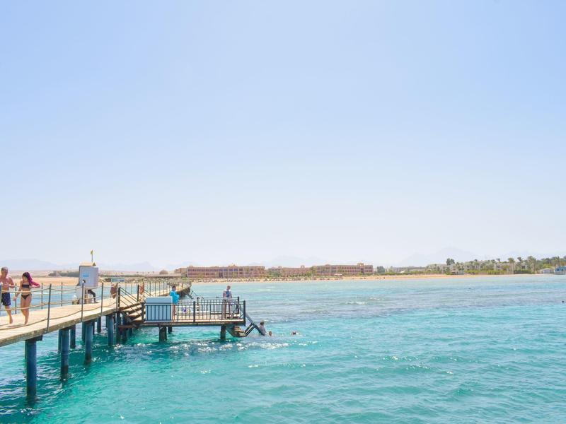 Holzsteg mit Menschen und klarem türkisfarbenem Wasser unter blauem Himmel am Strand.