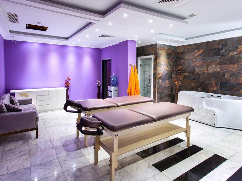 Ein Raum mit violetten Wänden, weißer Decke, massiertisch in der Mitte und Sofa an der Wand.