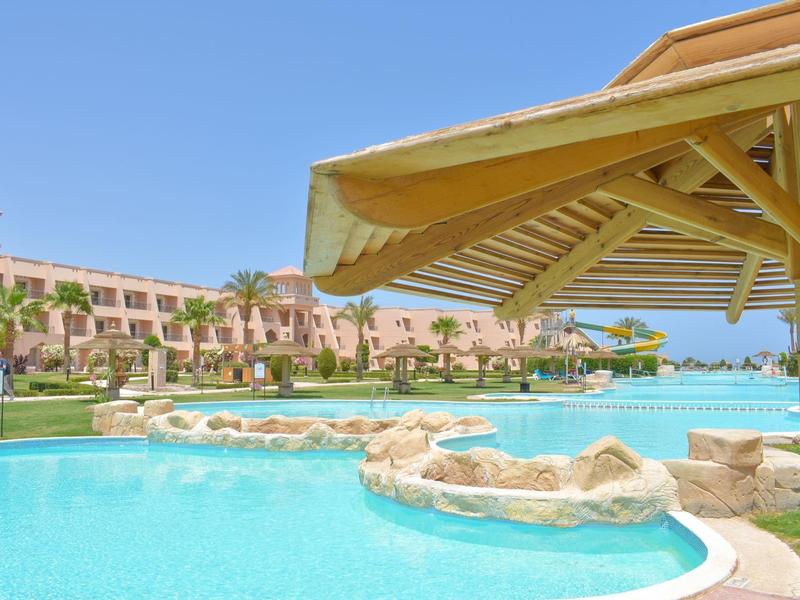 Großer Außenpool mit Felsen, Sonnenschirmen und rosa Gebäuden unter blauem Himmel.