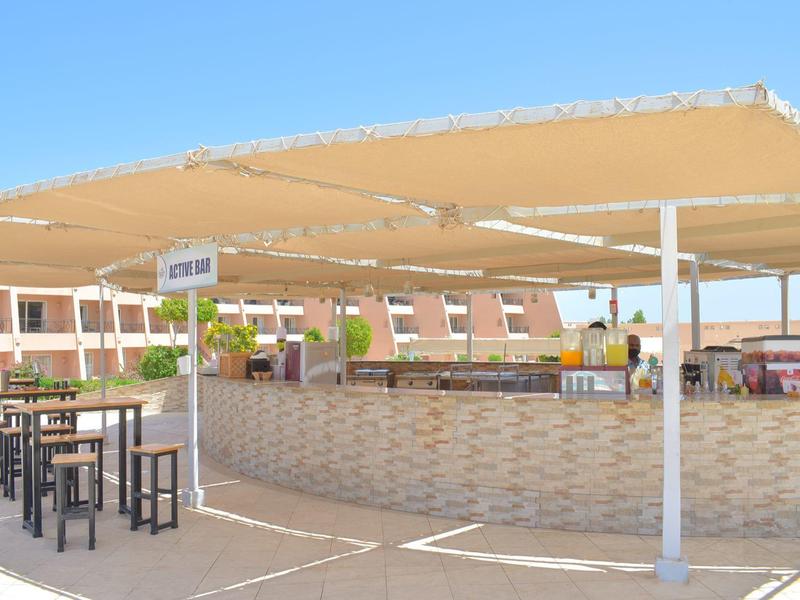 Outdoor-Barbereich mit Tresen, Hockern und Sonnendach vor rotem Gebäude bei klarem Himmel.