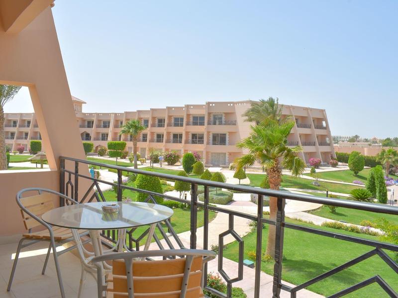 Balkon mit Metallmöbeln, Palmen und Blick auf gepflegte Grünflächen und großes Gebäude im Hintergrund.
