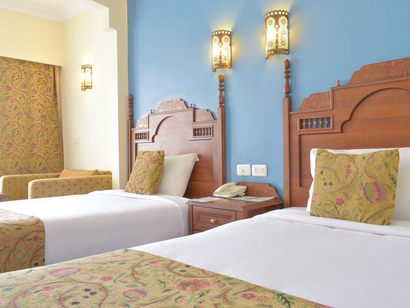 Hotelzimmer mit zwei Einzelbetten, braunen Holzbetten und blauer Wand, dekorative Lampen und Musterstoffe.